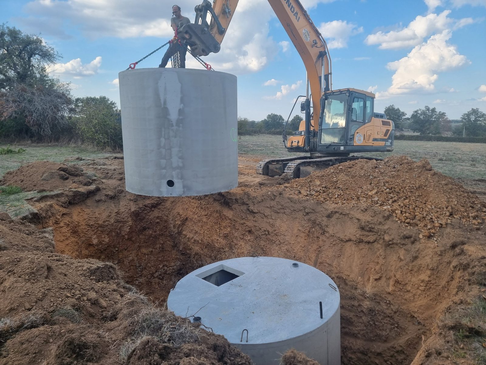 Chantier LOWASIS : une pelle mécanique descend un anneau de cuve en béton dans une fouille, à côté d'une première cuve déjà posée