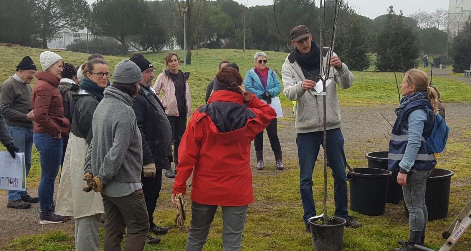 Chantier participatif de plantation : un groupe d'habitants rassemblés autour d'un jeune arbre à mettre en terre, sur une prairie.
